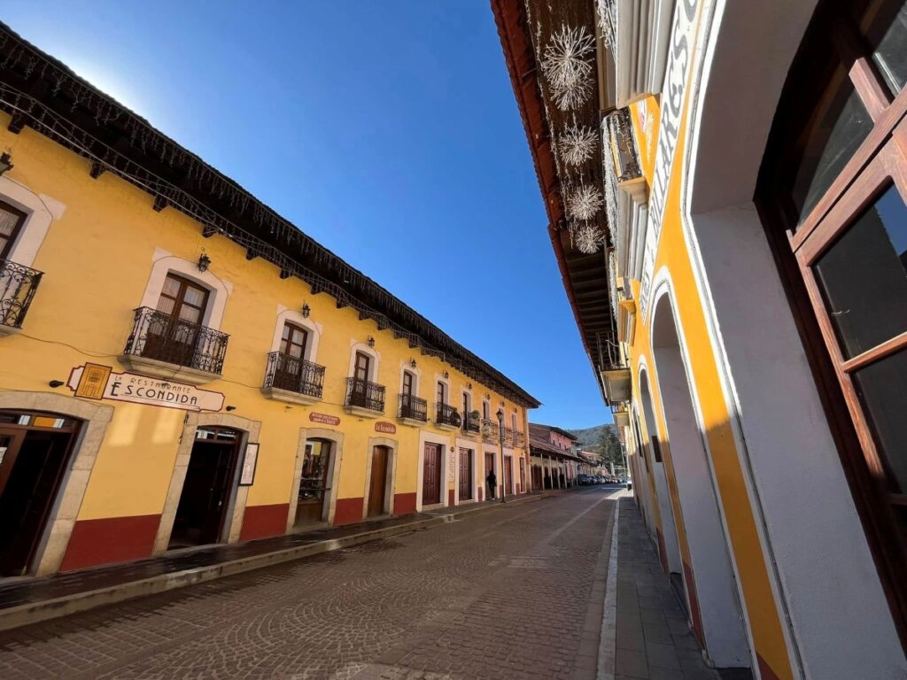 Calle empedrada en un Pueblo Mágico de Hidalgo, flanqueada por casas coloniales de fachada amarilla con balcones de hierro y cielo despejado.