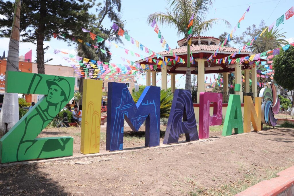 Coloridas letras de Zimapán en la plaza principal, decoradas con motivos regionales y rodeadas de banderines festivos, con el kiosco al fondo.