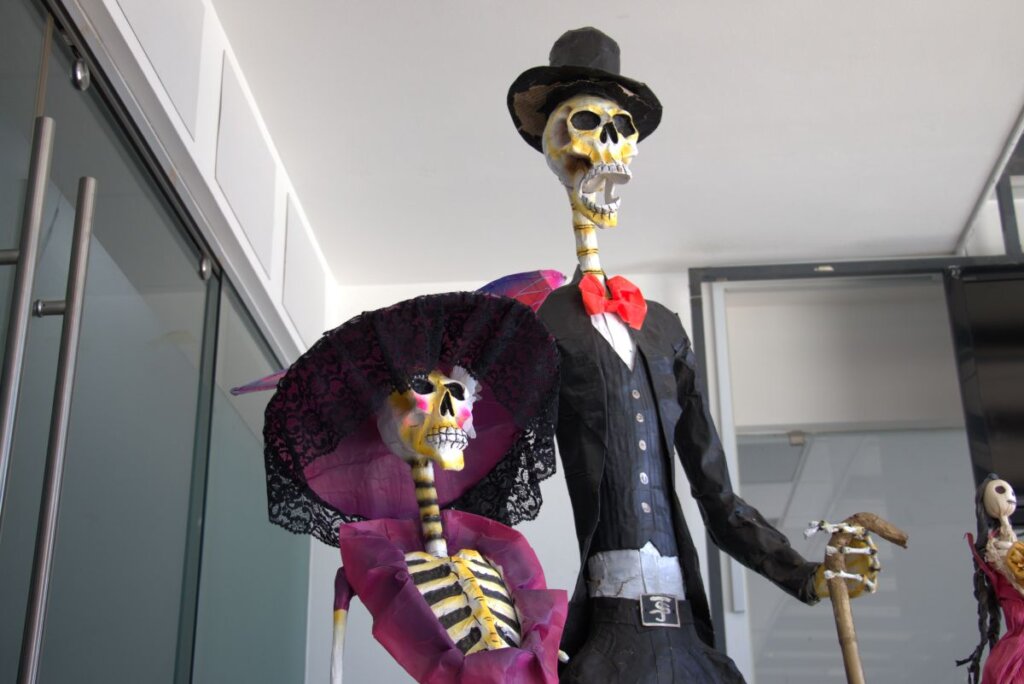 Dos figuras de papel maché representan a una pareja de esqueletos: él con sombrero y moño rojo, ella con un gran sombrero negro con encaje y vestido morado. Ambas esculturas están colocadas frente a una pared de cristal, como parte de las decoraciones del Día de Muertos en Tepeapulco.