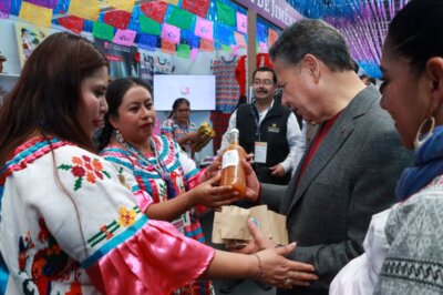 En la imagen, una mujer con vestimenta tradicional muestra al gobernador de Hidalgo una botella artesanal de bebida típica durante su recorrido por los pabellones del Tianguis Nacional de Pueblos Mágicos 2025 en Pachuca. A su alrededor se observan otras expositoras con trajes bordados y coloridos adornos de papel picado decorando el stand.