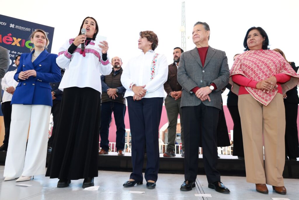En la imagen se observa a varias personas sobre un escenario durante la inauguración del Tianguis Nacional de Pueblos Mágicos 2025 en Pachuca. Una de las participantes sostiene un micrófono mientras dirige un mensaje al público. A su alrededor, autoridades federales, estatales y municipales escuchan atentamente el discurso, en un ambiente institucional y de celebración cultural.