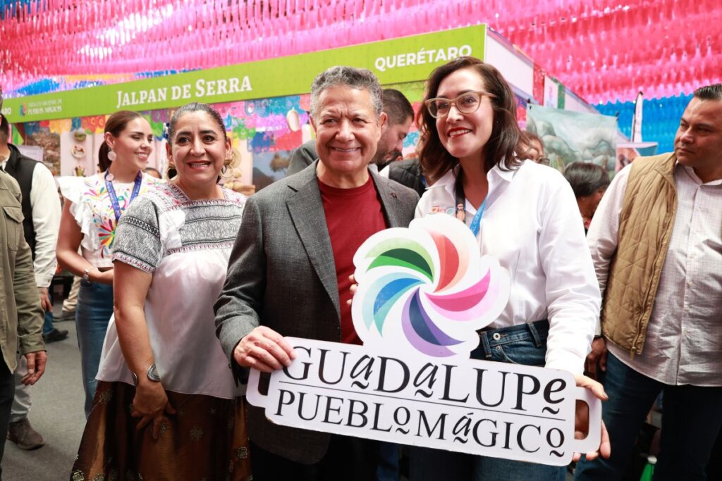 En la imagen se observa a varias personas sonriendo durante el recorrido por los pabellones del Tianguis Nacional de Pueblos Mágicos 2025 en Pachuca. Una mujer sostiene un cartel con el logotipo de “Guadalupe, Pueblo Mágico”, mientras al fondo se aprecian los stands de otros municipios como Jalpan de Serra, Querétaro, decorados con coloridos adornos de papel picado.