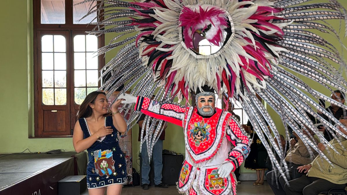 Danzante del Carnaval de Tlaxcala con traje tradicional y penacho de plumas coloridas baila junto a una joven durante una presentación cultural en un recinto histórico.