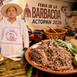 Elvis González, finalista más joven de la Feria de la Barbacoa Actopan 2026, aparece junto a una mesa con barbacoa tradicional, mientras al fondo se aprecia el ambiente festivo del concurso gastronómico en Actopan.