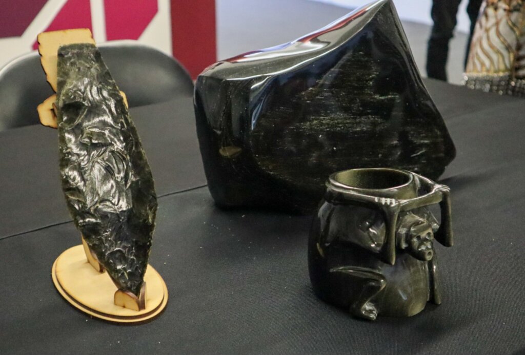 Piezas artesanales elaboradas en obsidiana negra, entre ellas una punta de lanza tallada, un bloque de roca volcánica y una taza con relieve de rostro prehispánico, exhibidas durante la presentación del Festival de la Obsidiana Dorada 2026 en Nopalillo, Epazoyucan.