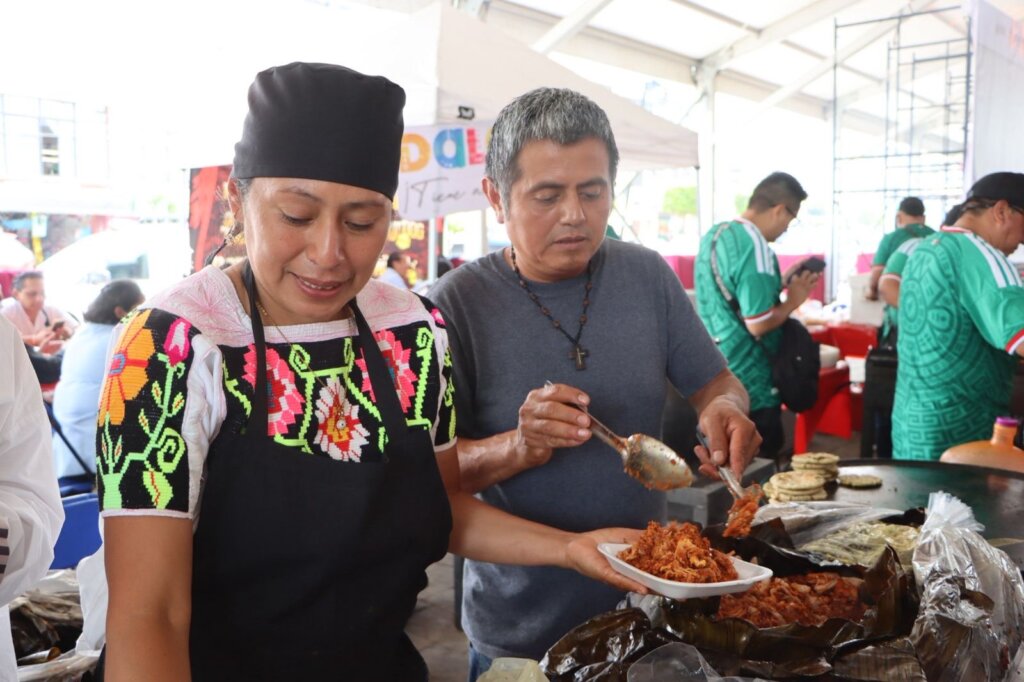 Cocinera tradicional sirve un platillo típico hidalguense durante una muestra gastronómica, acompañada de un expositor, en un encuentro que promueve la cocina regional y los sabores de los Pueblos con Sabor de Hidalgo.