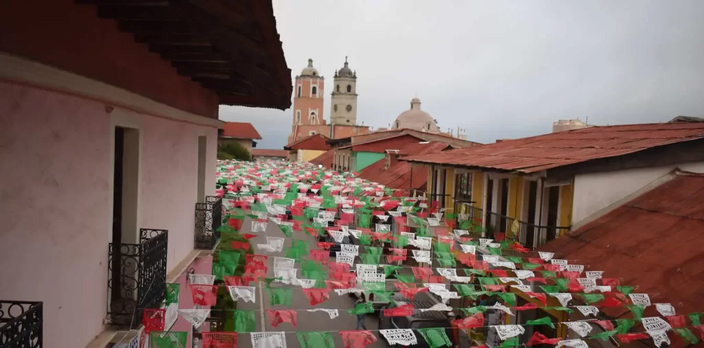 Calles de Real del Monte decoradas con papel picado verde, blanco y rojo con la Parroquia de Nuestra Señora de la Asunción al fondo durante las Fiestas Patrias.