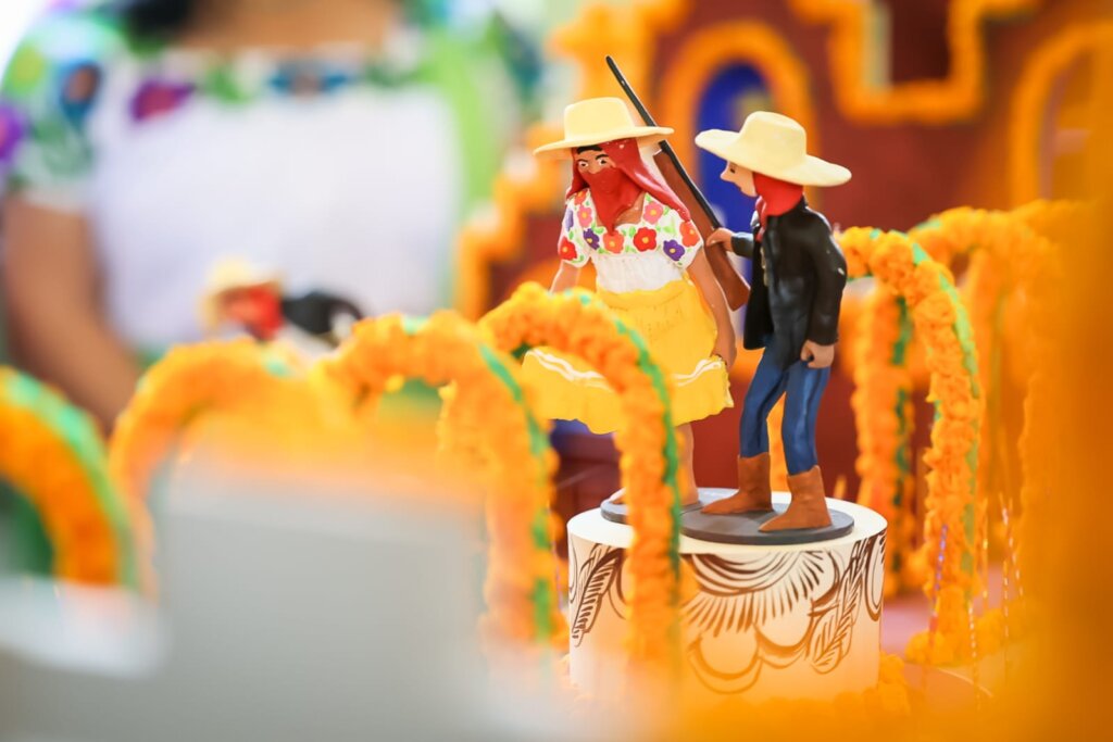Figuras de dos danzantes con sombreros y pañuelos rojos entre arcos florales anaranjados en la maqueta de Xantolo 2026.