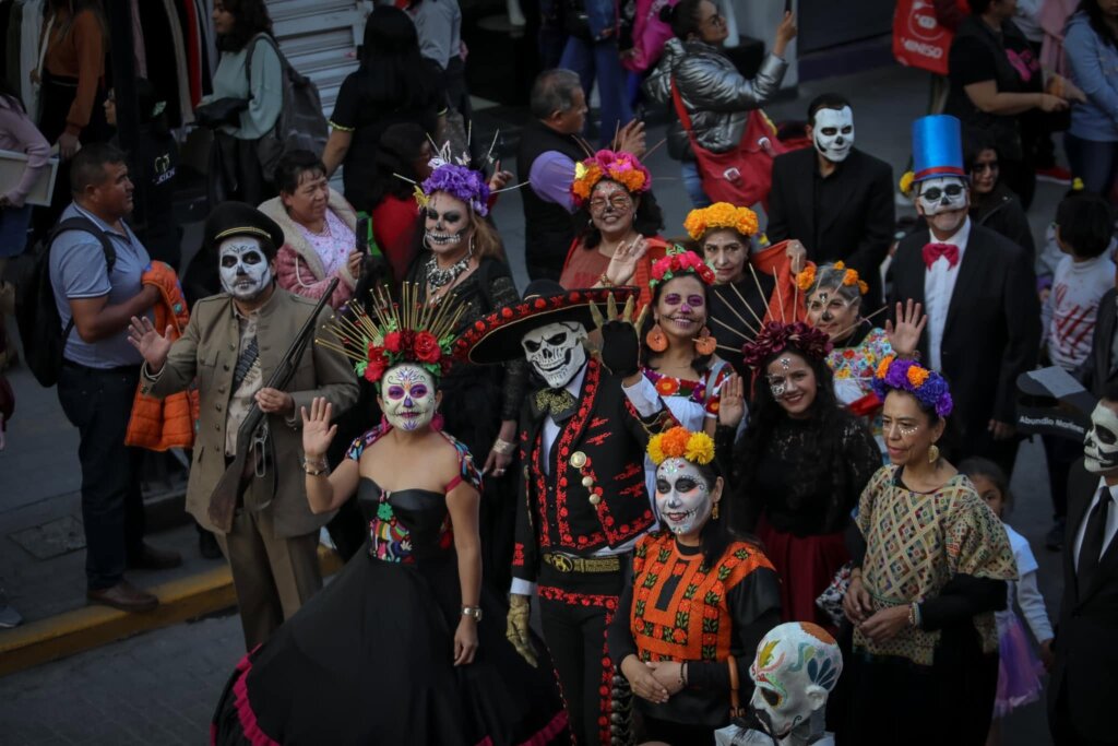 Grupo de participantes caracterizados como catrinas y catrines, con maquillaje de calaveras, trajes tradicionales y coronas de flores, durante un desfile de Día de Muertos en las calles de Pachuca, Hidalgo.