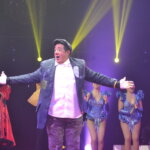 Payaso con gorra y saco de parches abre los brazos frente al elenco de Vie de Cirque, bajo luces amarillas y violetas